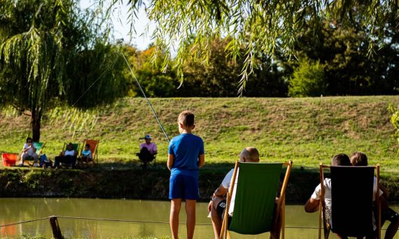 Young boy fishing by a pond on a sunny day, surrounded by trees and relaxed people.