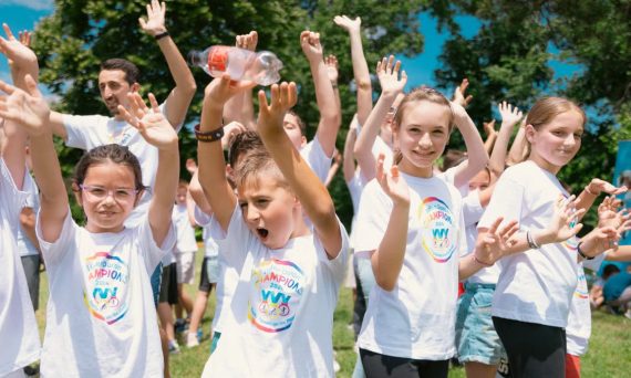 Group of cheerful children raising hands in a park during a sunny day, wearing t-shirts with event logos.